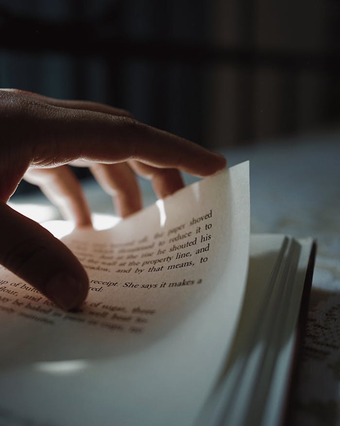 about-us Close-up of a hand turning a page in an open book with blurry text in dim light.