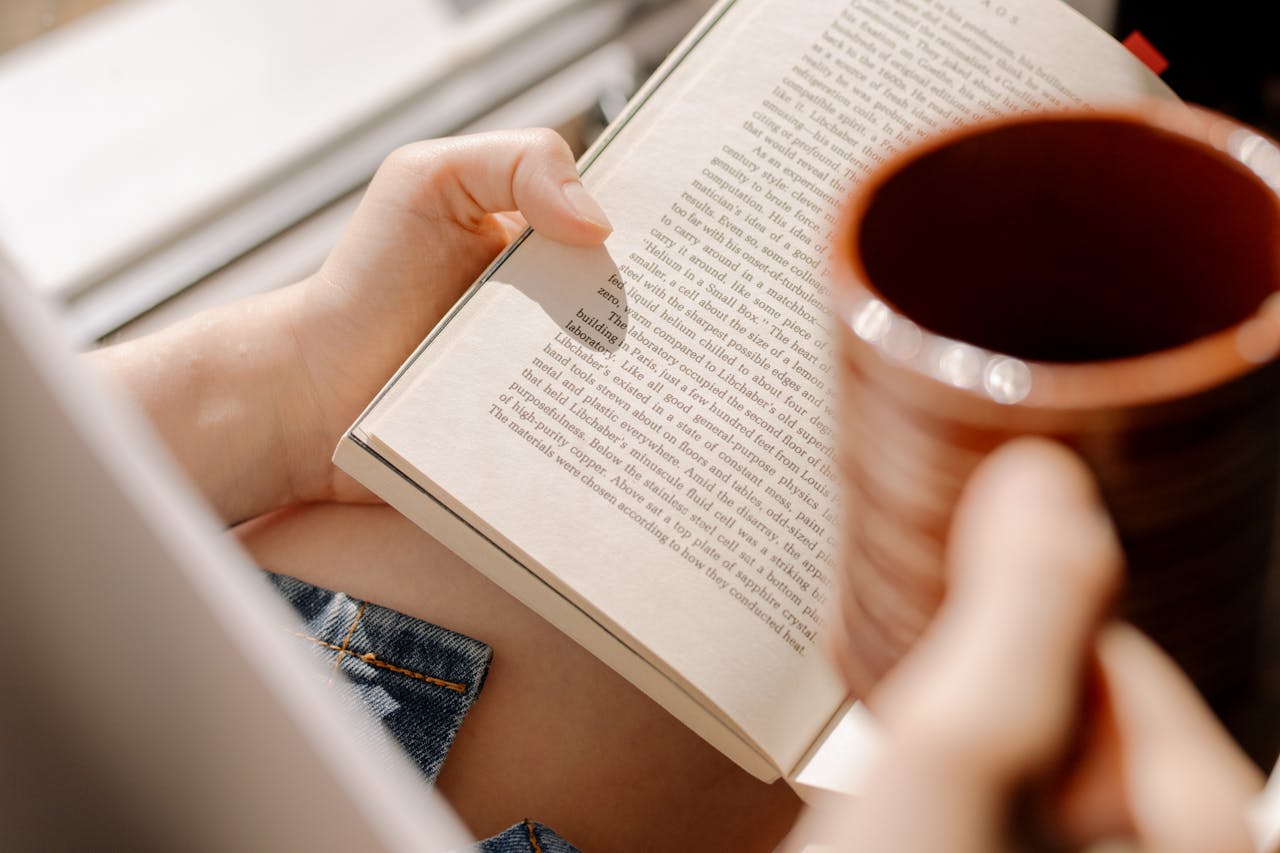 A person reading a book by the window, enjoying a warm cup of tea.