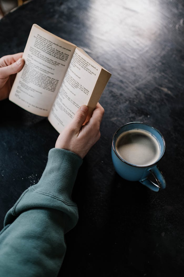Close-up of hands holding a book next to a ceramic cup of coffee on a dark table.